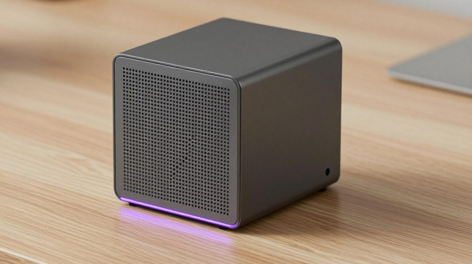 A compact graphite AiServa AI server with a thin violet light along its base on a pale oak desk, a laptop softly out of focus behind it