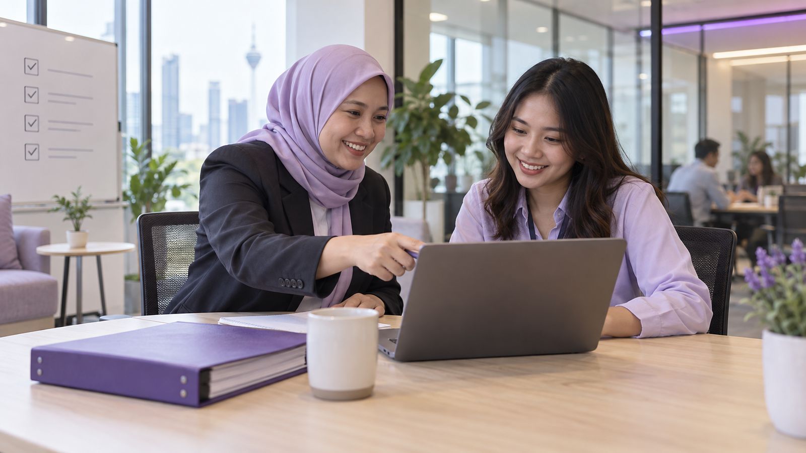 A Malaysian HR manager in a bright meeting room guiding a new employee through an onboarding checklist on a laptop, an employee handbook on the table