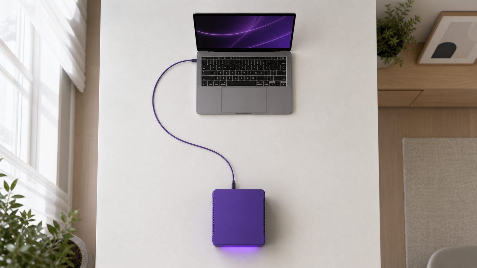 Overhead view of a laptop and a violet local AI server on a desk joined by a single neatly routed cable